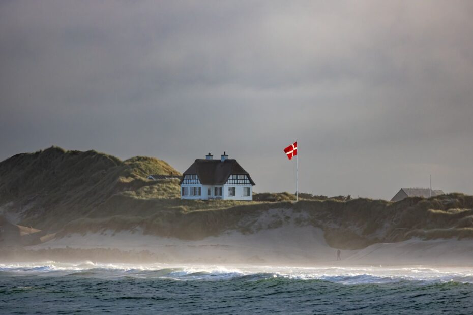 Quaint cottage by Danish coastline with waving flag and rolling waves.
